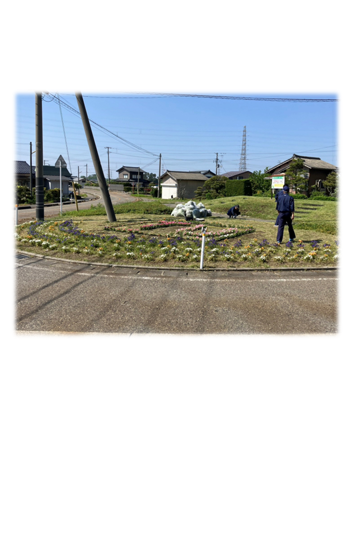 色とりどりの花が植えられた道路沿いの広場の一角で従業員が3名、除草作業している写真。