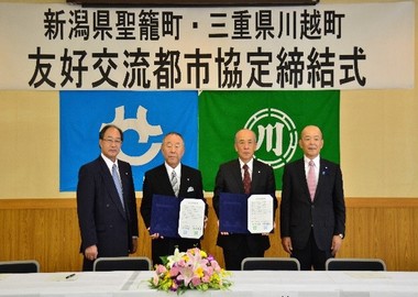 「新潟県聖篭町・三重県川越町友好交流都市協定締結式」と書かれた横断幕と聖籠町と川越町の町章旗が設置された壁の前で4名の関係者が締結書を持ち記念撮影を行っている写真