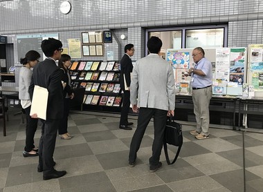 掲示板に貼られた資料の前で関係者が集まり話をしている様子の写真